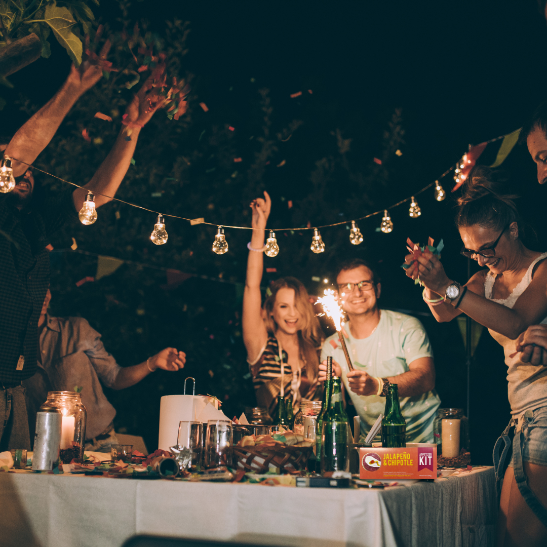 People celebrating outdoors with sparklers and drinks under string lights with a Jalapeno & Chipotle appetizer kit on the table.