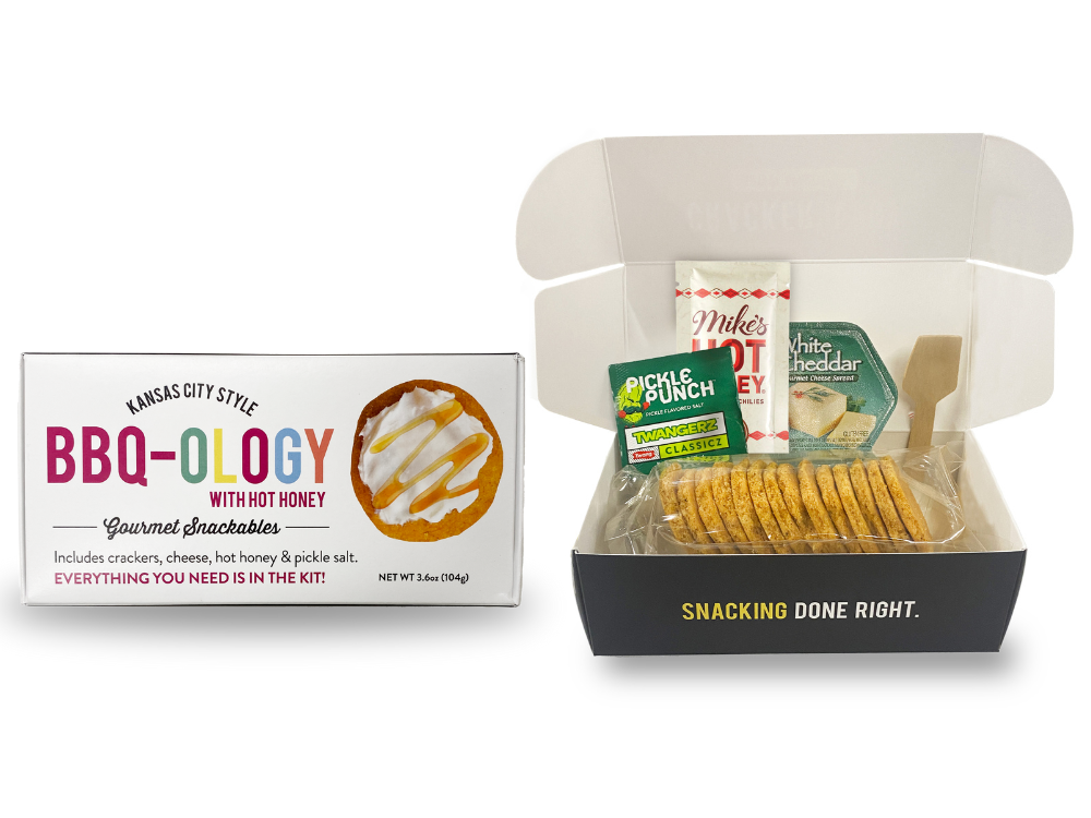 An open snack box labeled 'Snackable BBQ-OLOGY Crackerology Kit', displaying BBQ crackers, cheese, pickle salt, and hot honey inside.