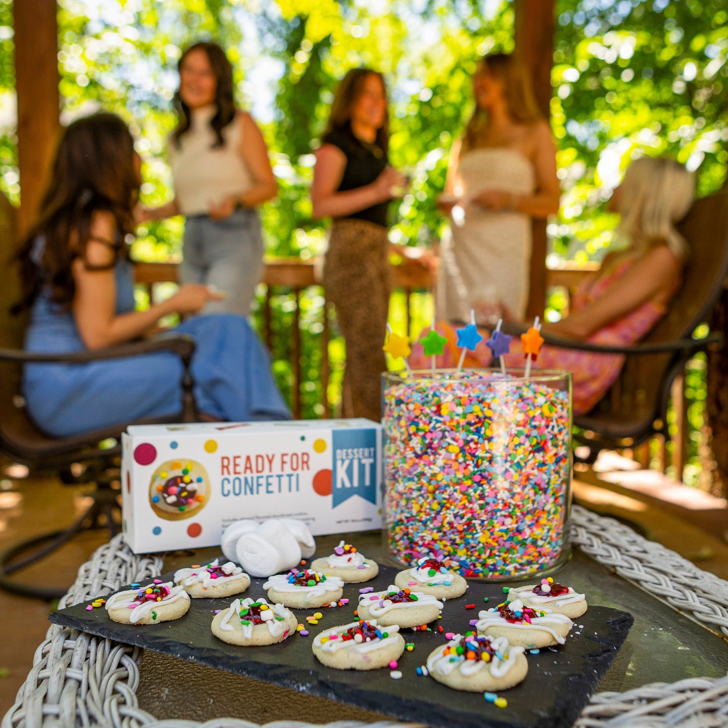 Table with cake confetti cookies supplies and treats outdoors, people in the background