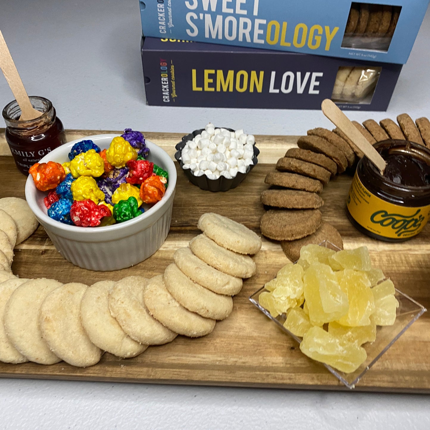 Wooden tray with lemon and s'more cookies, candies, and a jar of jam on a white background